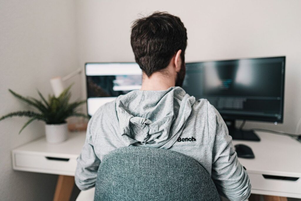 Person in a light gray "Bench" hoodie working at a dual-monitor computer setup in a modern home office, with a potted plant on the desk and dark-themed screens displaying code.