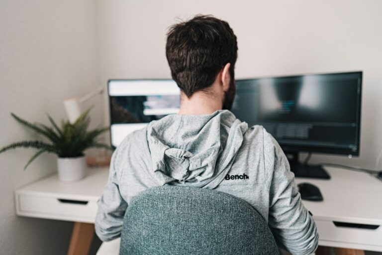 Person in a light gray "Bench" hoodie working at a dual-monitor computer setup in a modern home office, with a potted plant on the desk and dark-themed screens displaying code.